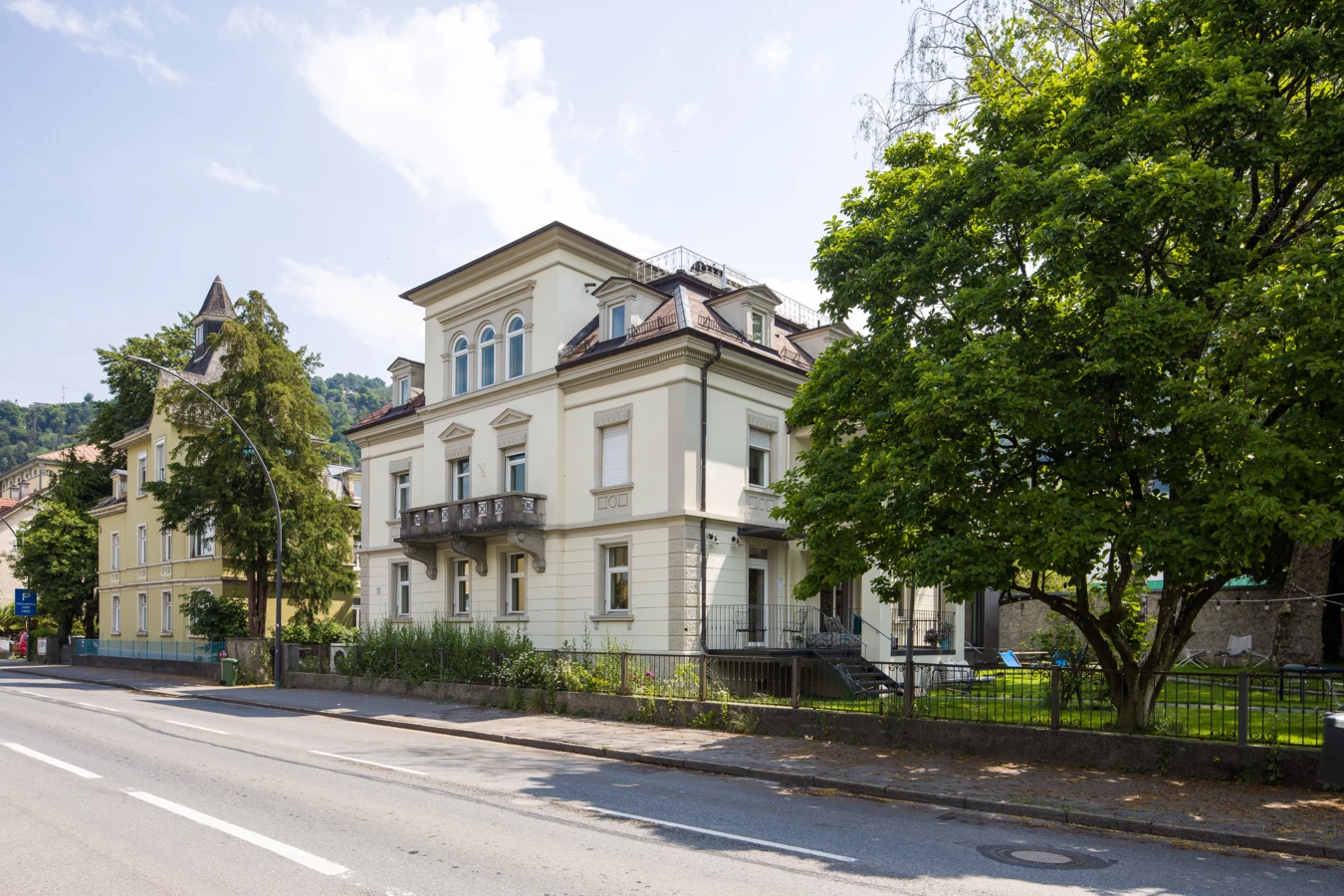 Eine Straße mit einem großen, cremefarbenen Gebäude mit Balkonen und einem hohen Baum daneben (Wunderkind, Backstube).