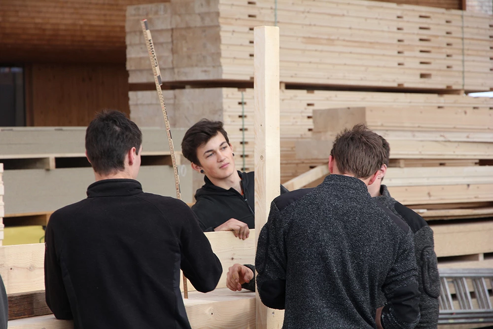 Drei junge Männer arbeiten mit Holzplatten und -balken in einer Halle, umgeben von gestapeltem Holzmaterial. Einer der Männer hält ein Maßband in der Hand (Lehrlingstag).