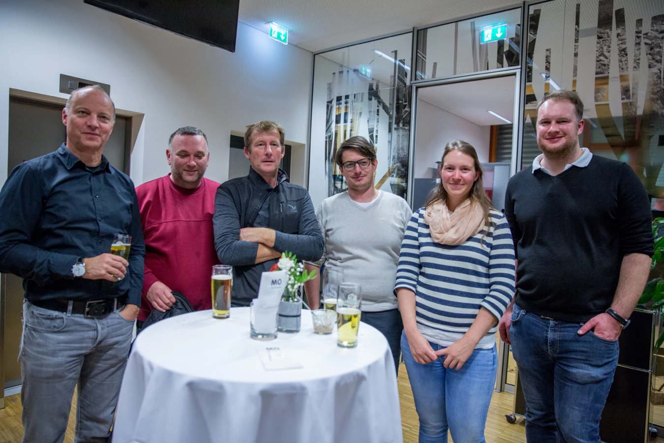 Eine Gruppe von sechs Personen, drei Frauen und drei Männern, stehen lächelnd bei einem Event mit Getränken auf einem Stehtisch (Hubert, Geburtstag).