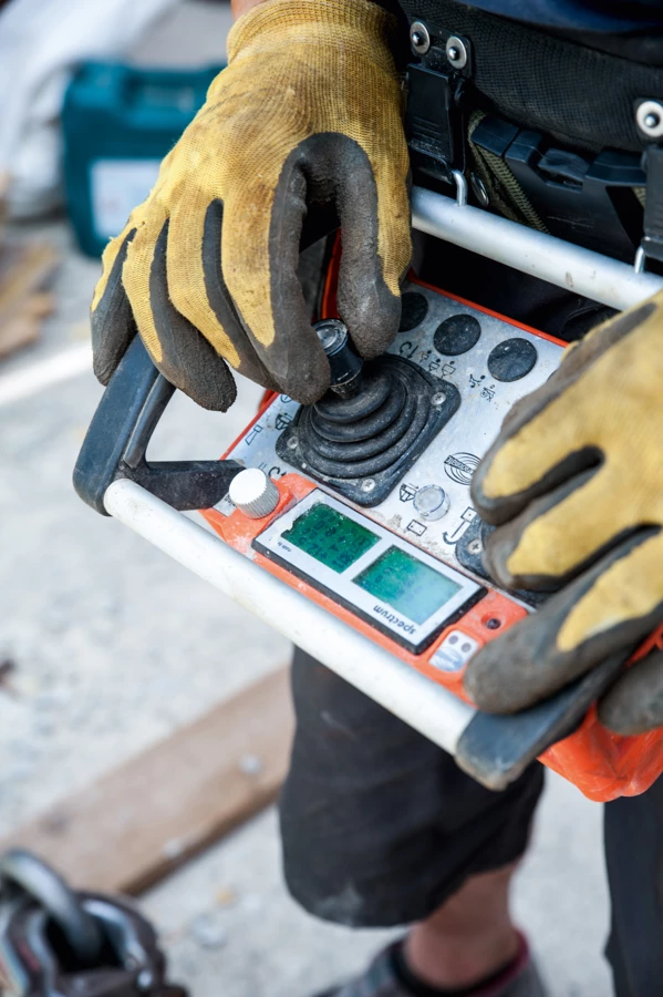 Arbeiter in gelben Schutzhandschuhen bedient ein orange-graues Funkfernsteuerungsgerät mit zwei Bildschirmen und einem Joystick (Baustelle, Hochbau)