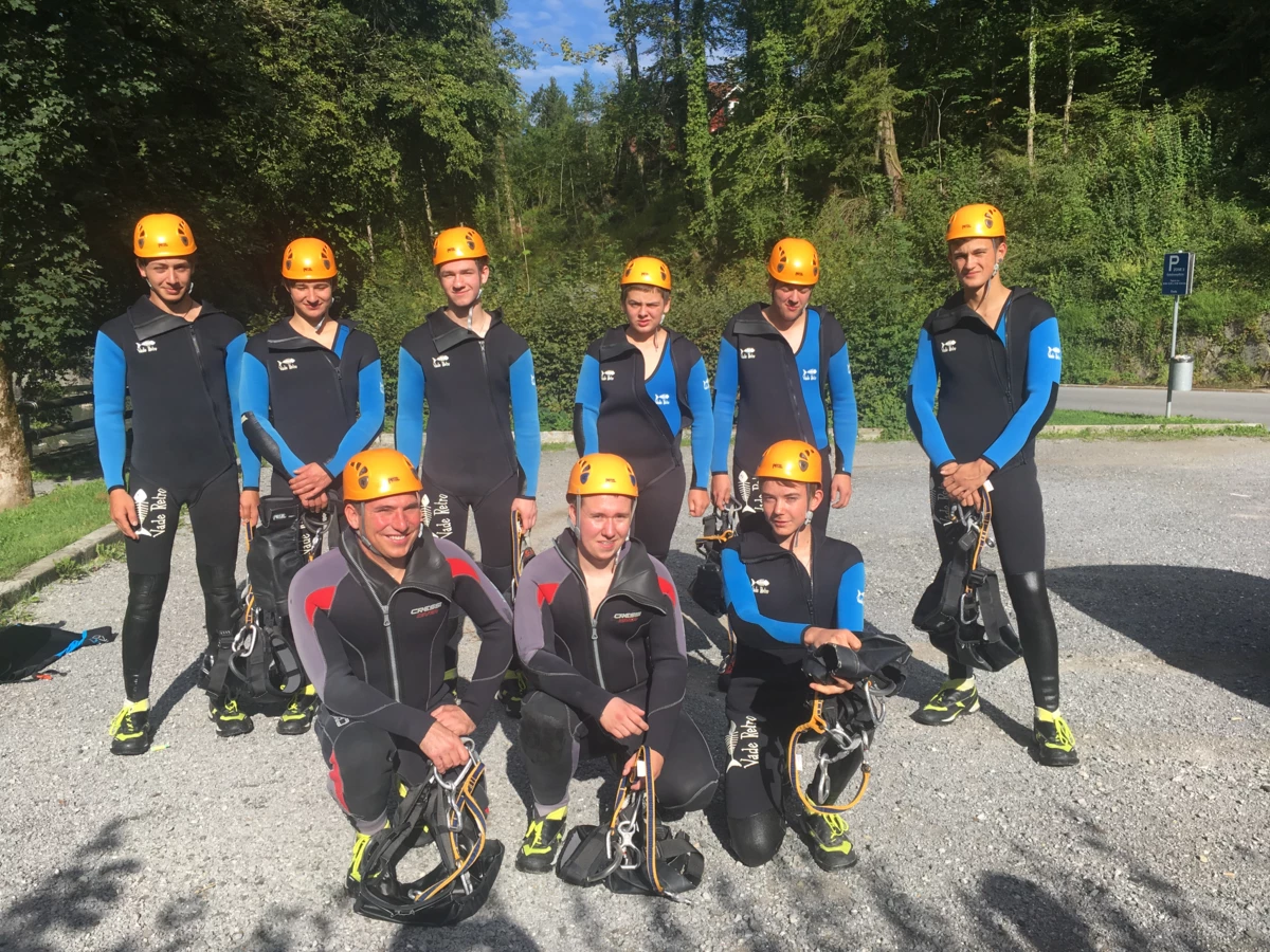 Eine Gruppe von neun jungen Erwachsenen in Neoprenanzügen und orangefarbenen Helmen, bereit für Outdoor-Aktivitäten (Lehrlingen, Rafting, Ausrüstung).
