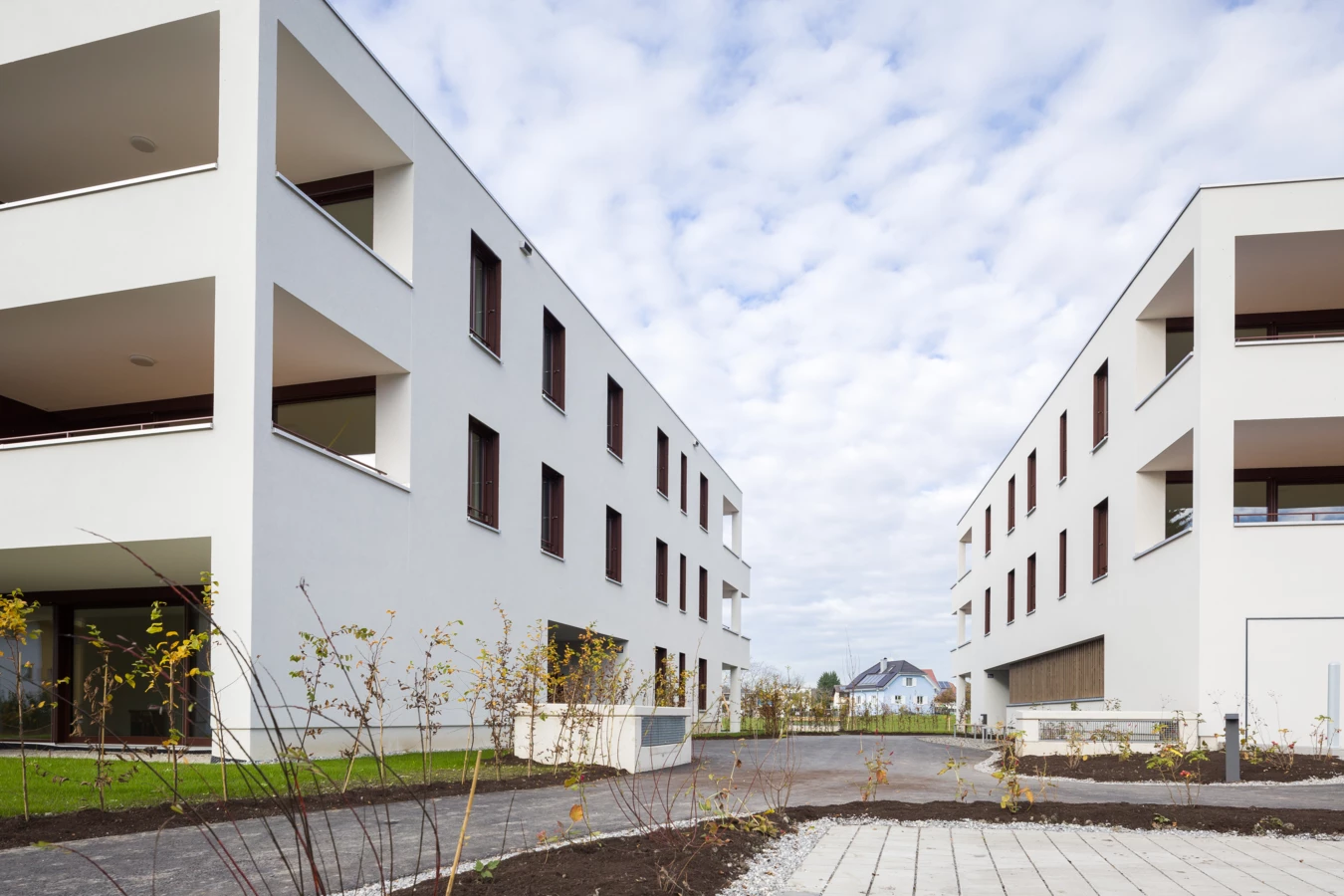 Eine Außenansicht zweier moderner, heller Wohngebäude mit Balkonen und braunen Fenstern, umgeben von junger Bepflanzung und einem befestigten Weg (Unterhochstegstrasse).