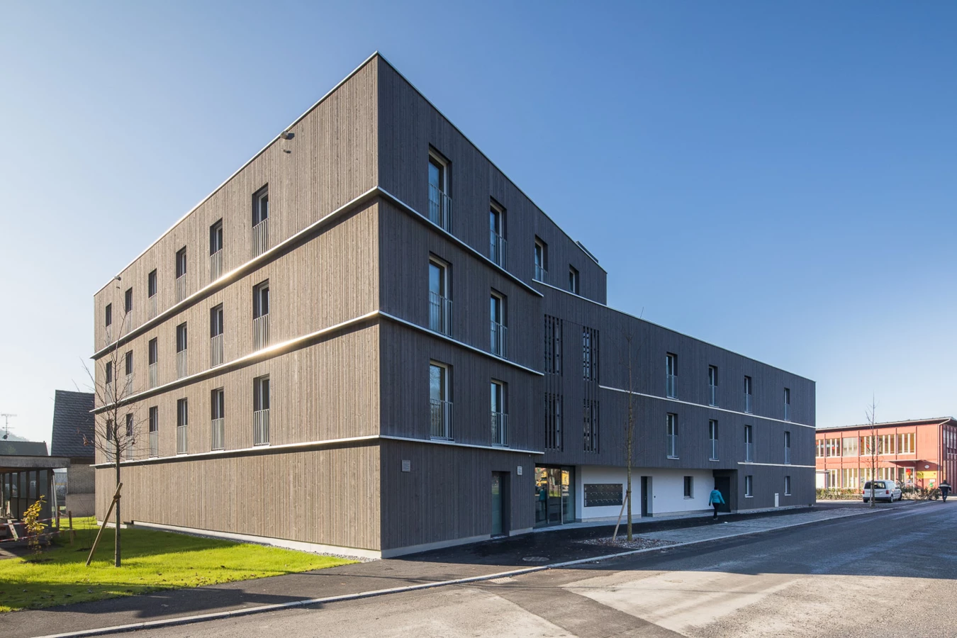 Modernes, mehrstöckiges Wohngebäude mit Holzfassade und Balkonen unter blauem Himmel, entlang einer Straße mit Grünflächen (Froschweg).