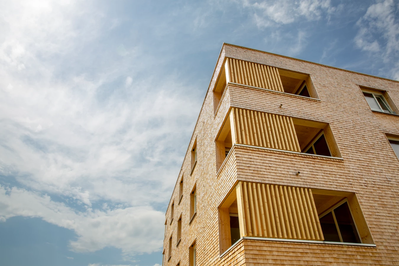 Holzfassade eines modernen Gebäudes mit vertikalen Holzlamellen und hellen Fenstern unter bewölktem Himmel (Hellbrunnenstraße).