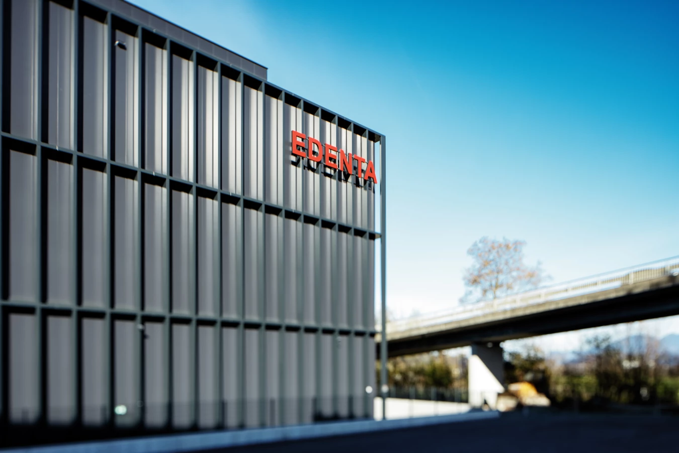 Modernes Gebäude mit dem roten Logo "EDENTA" an der Fassade unter blauem Himmel, einer Brücke und Bäumen im Hintergrund.