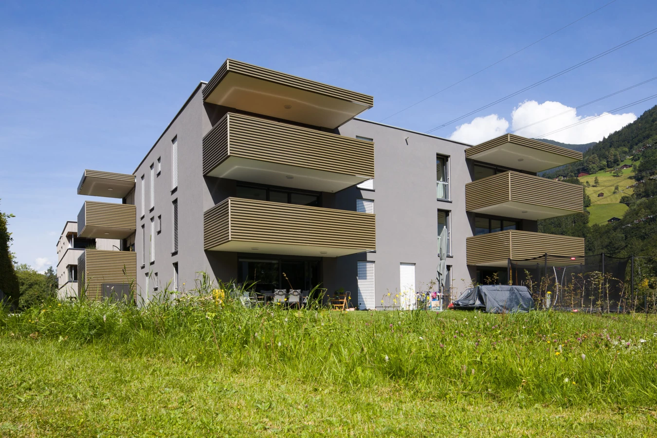 Modernes Wohnhaus in Vandans mit grauer Fassade, hellen Holzlamellenbalkonen auf einer grünen Wiese unter blauem Himmel (Bahnhofstraße, Wohnanlage, Bahnhofsstrasse).