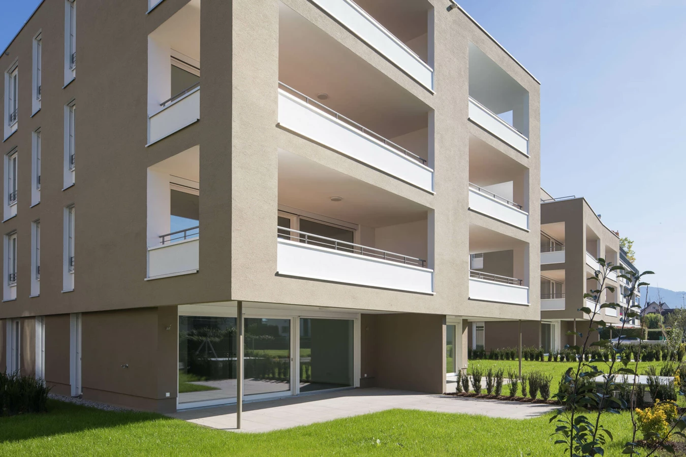 Moderne Wohnanlage Widum Lustenau mit beigefarbenen Fassaden, weißen Balkonen und Grünflächen unter blauem Himmel.