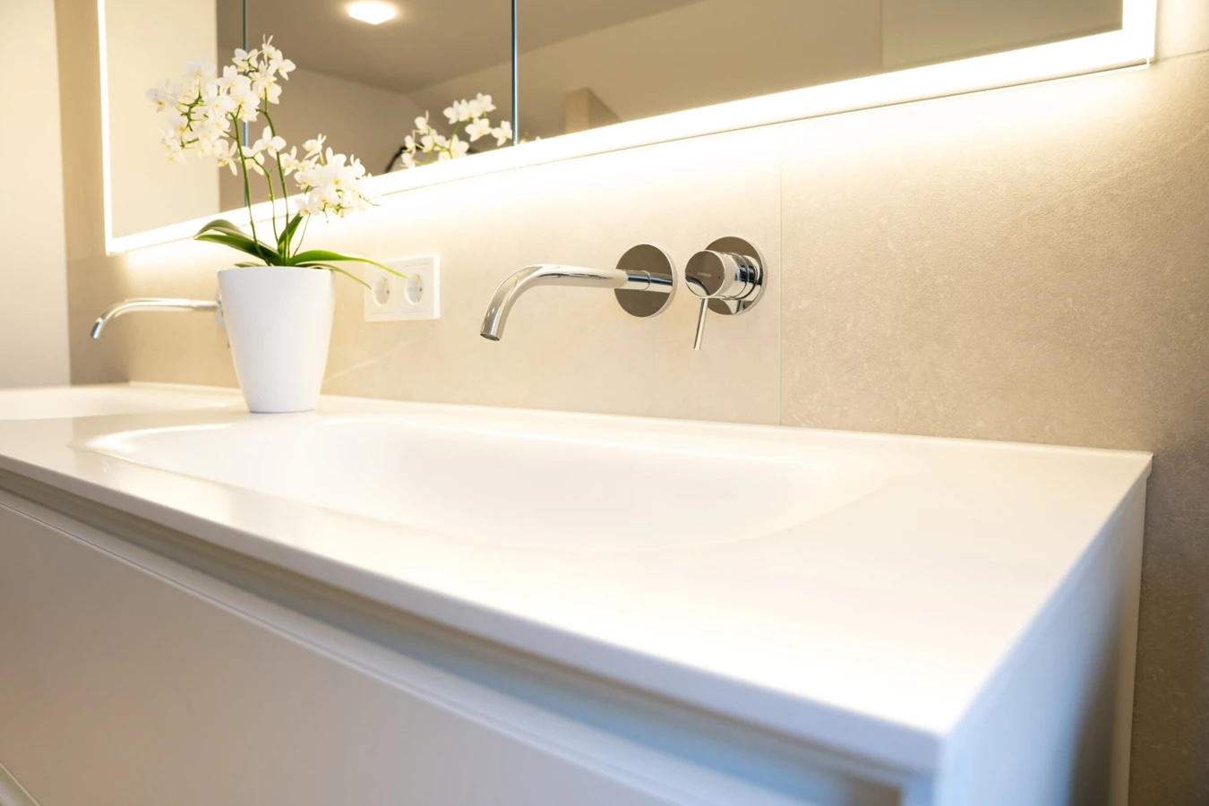 Modernes Badezimmer mit weißem Waschtisch, Wandarmatur, beleuchtetem Spiegel und einer weißen Orchidee in einem Topf (Generalsanierung, Bad).