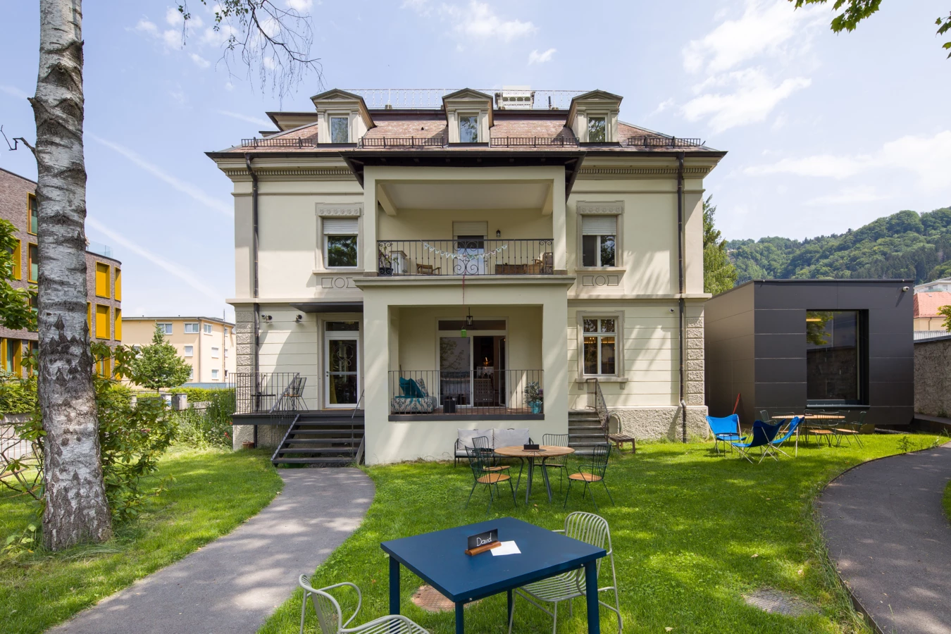Historisches helles Mehrfamilienhaus mit Vorgarten und Terrassen, ausgestattet mit Tischen und Stühlen auf grünem Rasen (Wunderkind, Backstube).