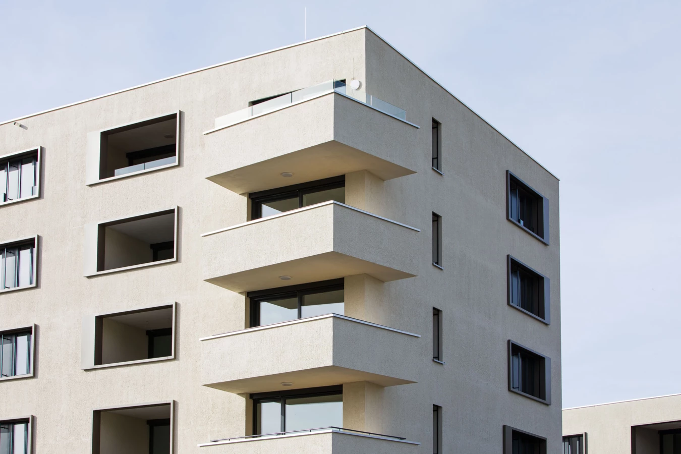 Moderne, hellbeige Fassade eines mehrstöckigen Wohngebäudes mit dunklen Fenstern und gestuften Balkonen an der Ecke (Bäumleareal).