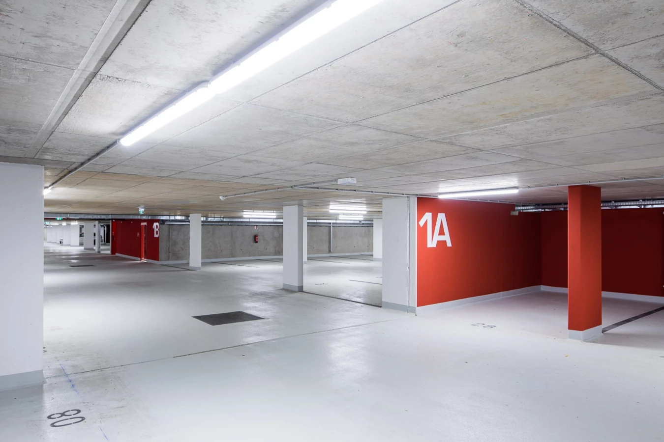 Leere Tiefgarage mit Betondecke, weiß gestrichenen Säulen und leuchtend roten Wänden, die mit den Zahlen 1A und 1B gekennzeichnet sind (Baeumlequartier).