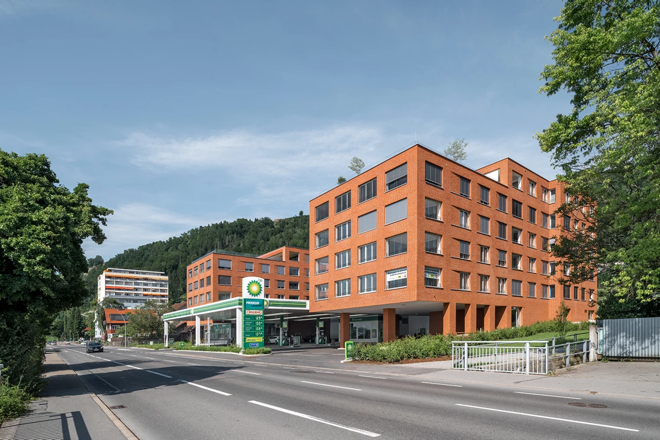 Zwei moderne orangefarbene Backsteingebäude neben einer BP-Tankstelle an einer Straße in Lochau, mit bewaldeten Hügeln im Hintergrund (Seeblick).