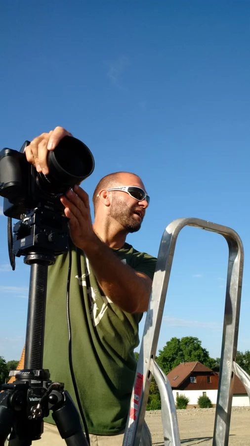 Mann mit Sonnenbrille richtet eine Kamera auf einem Stativ aus, hinter einer Leiter stehend, vor einem blauen Himmel.