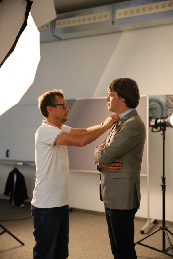 Ein Mann im weißen T-Shirt und Brille richtet dem anderen Mann im Anzug den Kragen bei einem Fotoshooting.