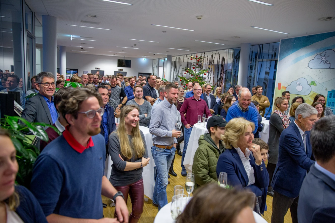 Eine große Gruppe lachender Menschen, die eine Präsentation oder Veranstaltung genießen, mit einem Weihnachtsbaum im Hintergrund (Hubert, Geburtstag).