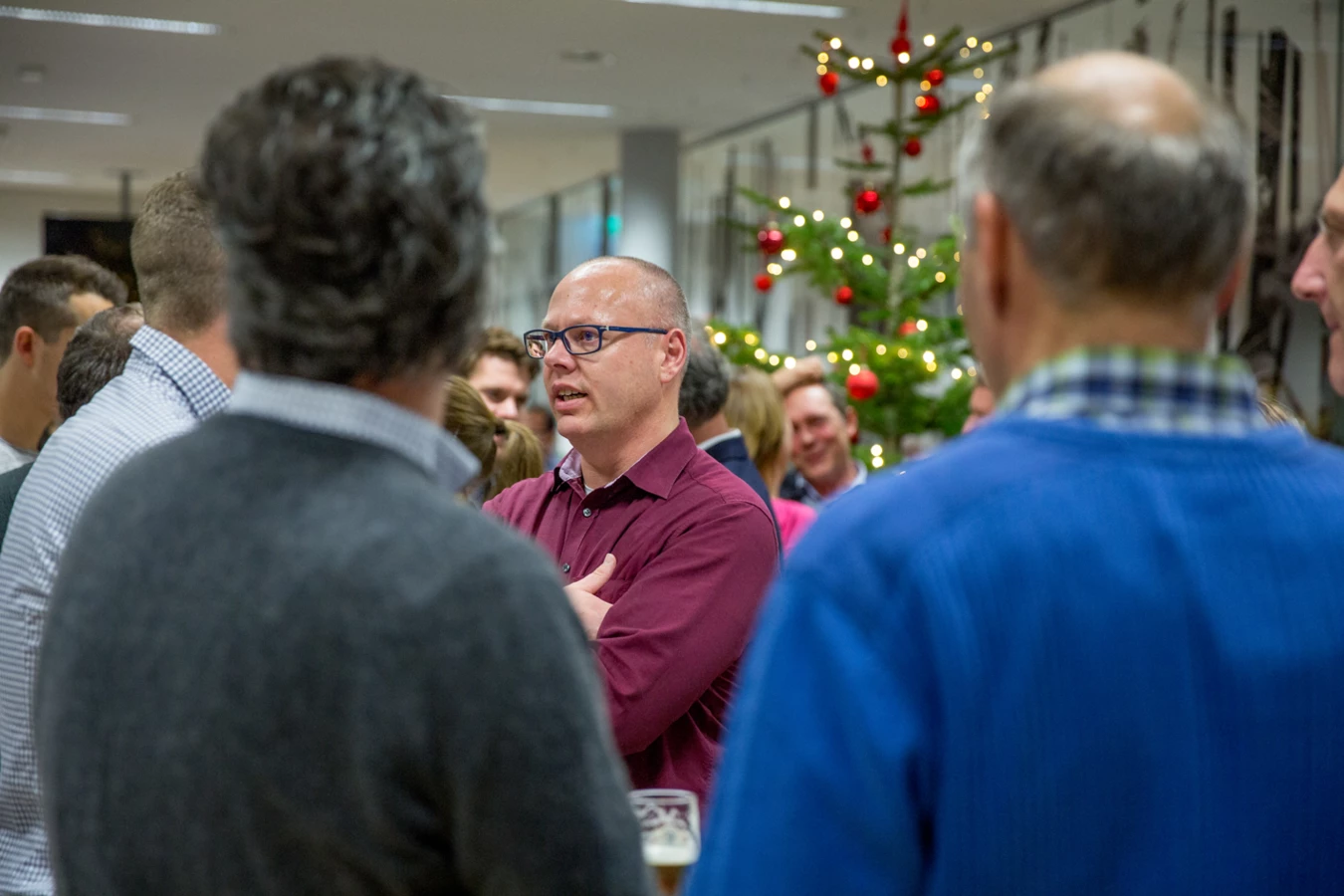 Ein Mann mit Brille und weinrotem Hemd spricht in einer Gruppe von Menschen, im Hintergrund ein Weihnachtsbaum (Hubert, Geburtstag).