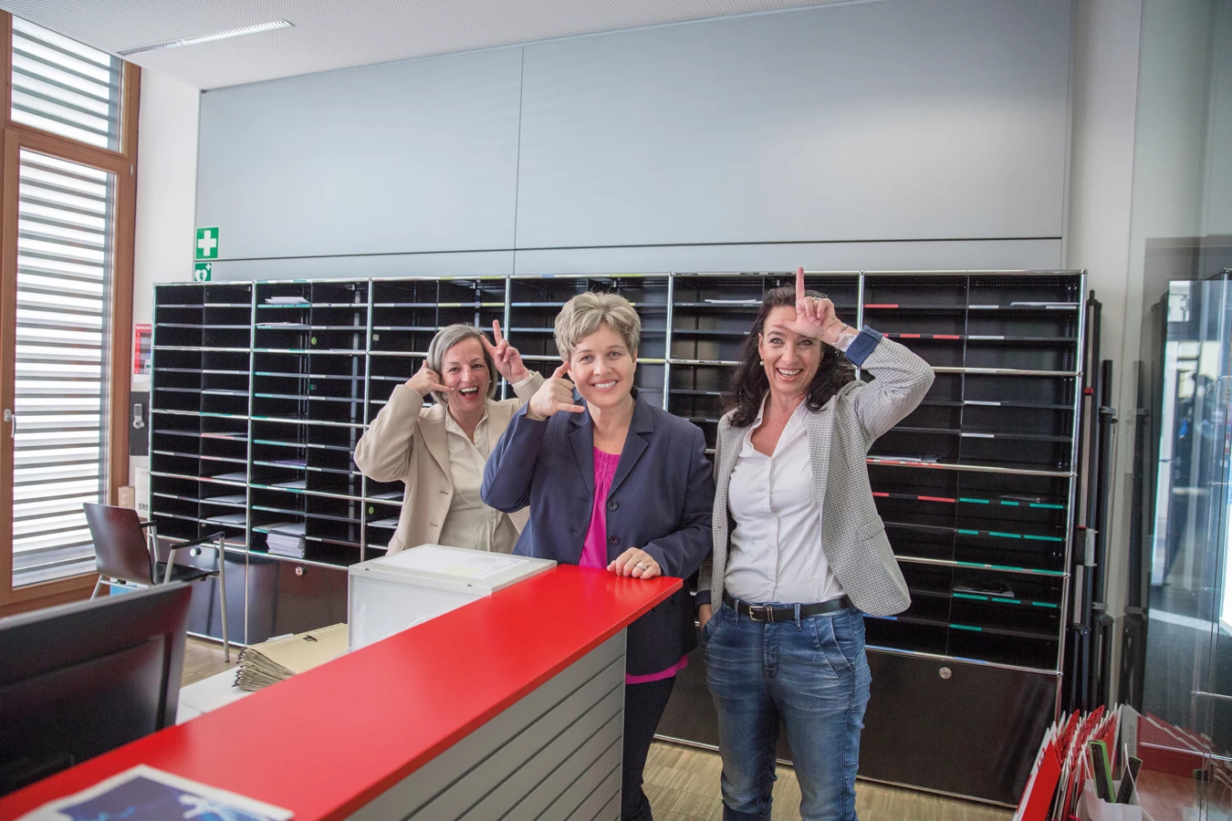Drei fröhliche Frauen posieren hinter einem roten Empfangstresen vor Briefregalen und einem Fenster (Empfangsdamen).