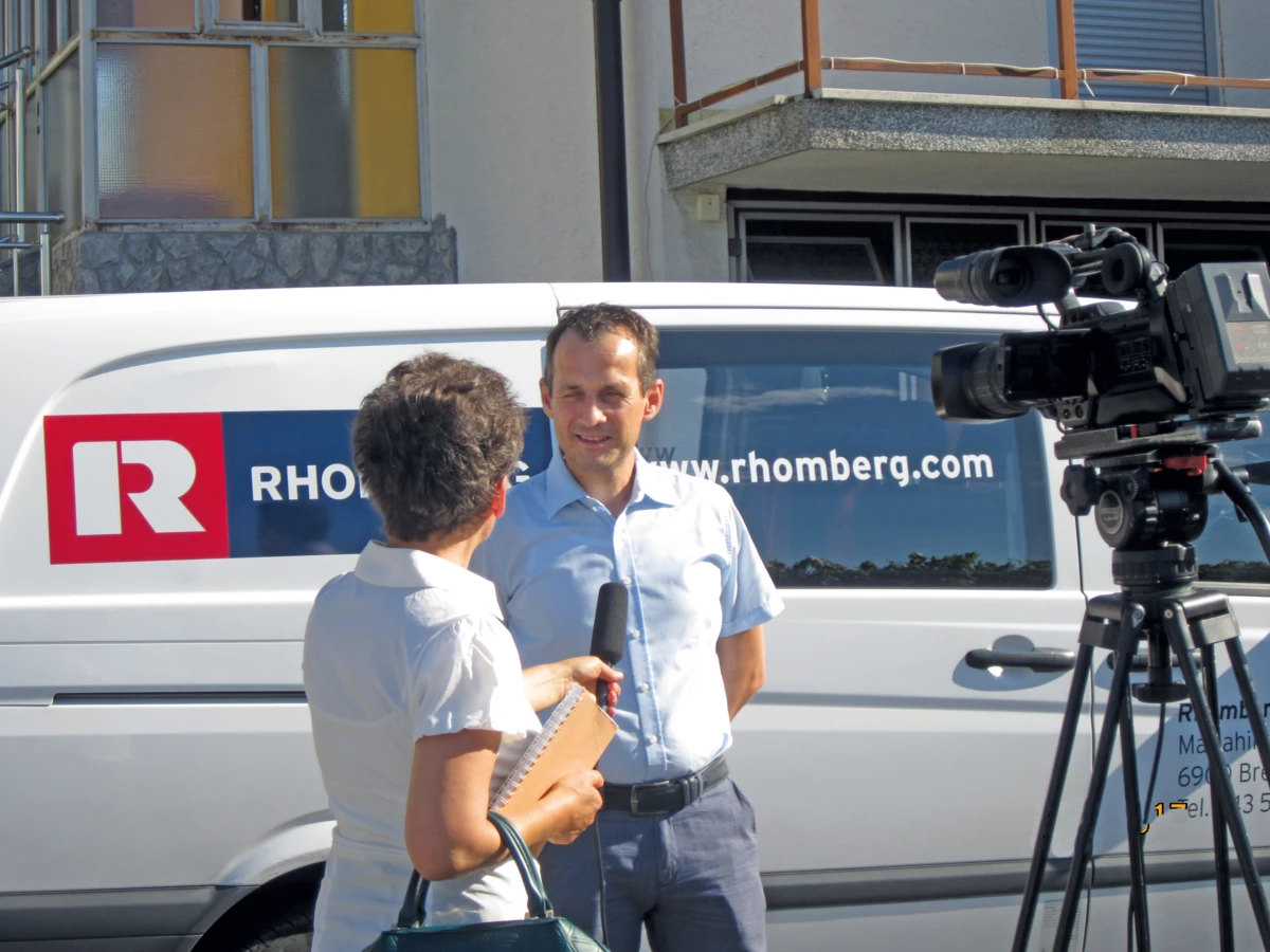 A man in a light blue shirt is interviewed by a woman holding a microphone and notebook, beside a Rhomberg Bau van and a camera on a tripod (pensioners)