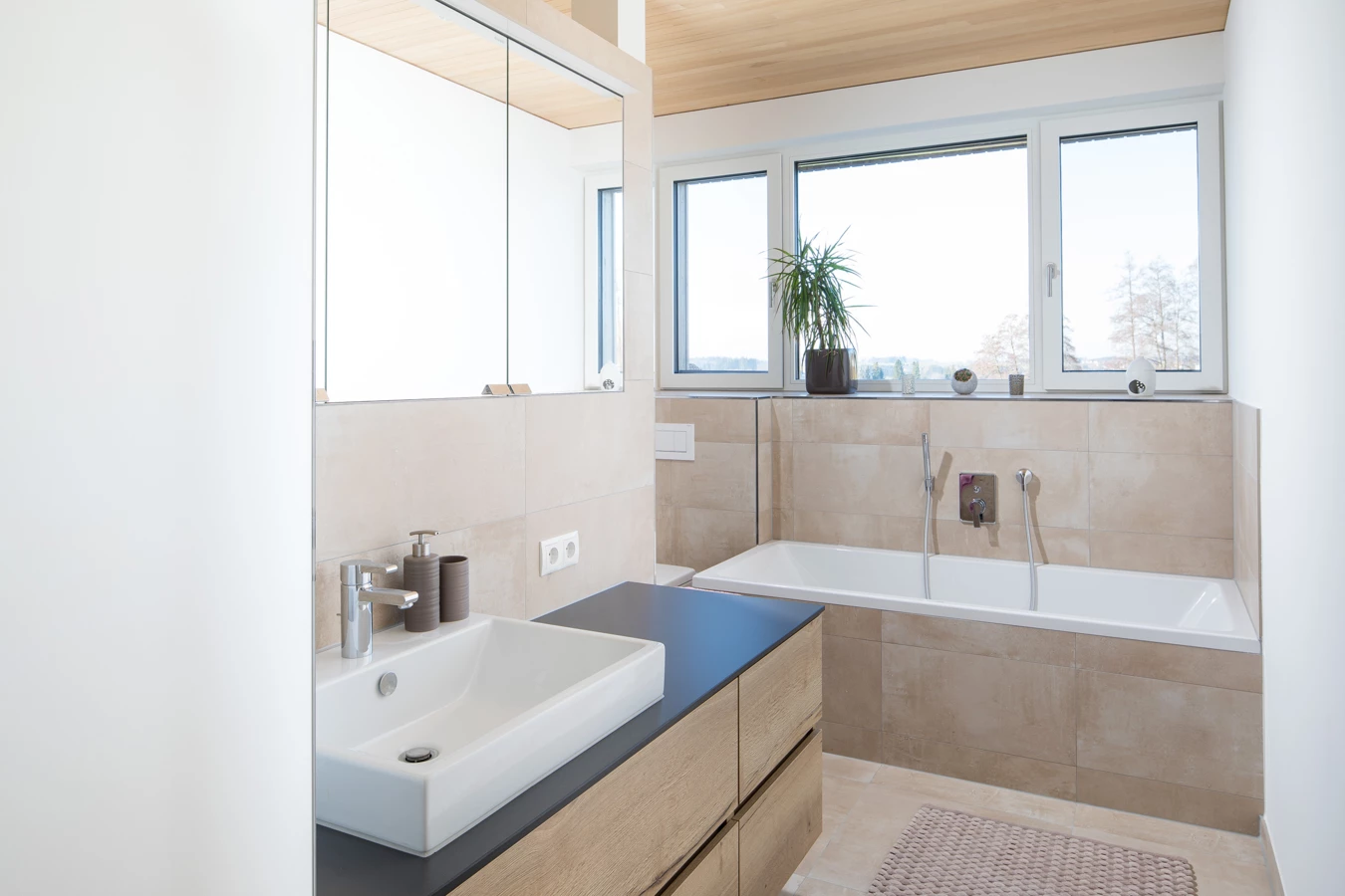 Modernes Badezimmer mit weißer Badewanne, Eckwaschbecken auf Holzunterschrank und einem großen Fenster (Hohenweiler)