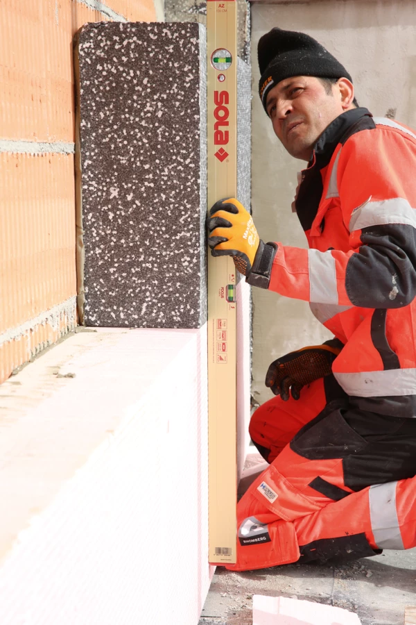 Arbeiter misst mit einer Wasserwaage die Ebene einer Dämmplatte an einer Backsteinwand (Verputzarbeiten, Hochbau).