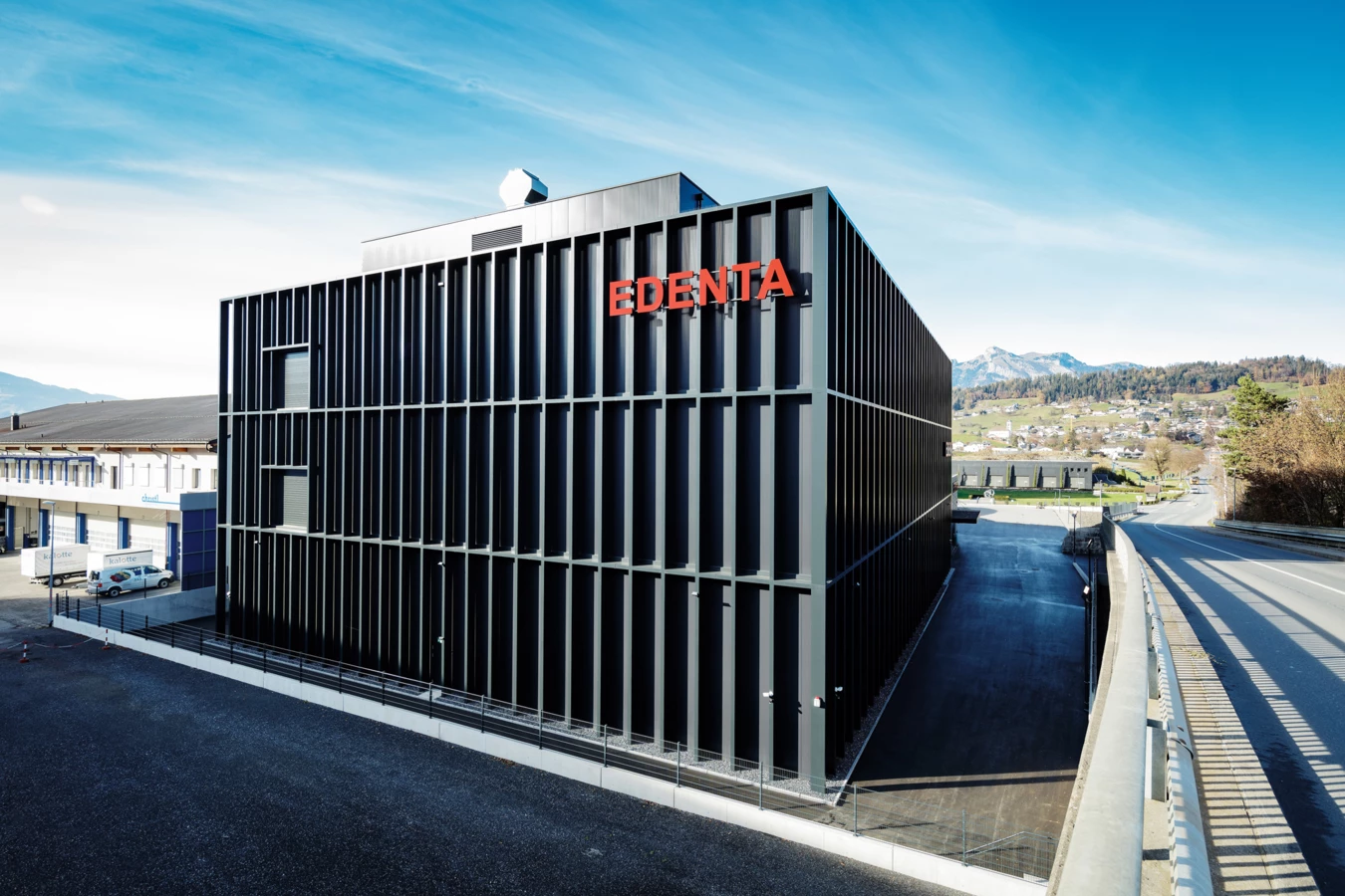 Modernes schwarzes Gebäude mit "EDENTA"-Schriftzug in Rot gegen einen blauen Himmel, daneben eine Straße und Berge.