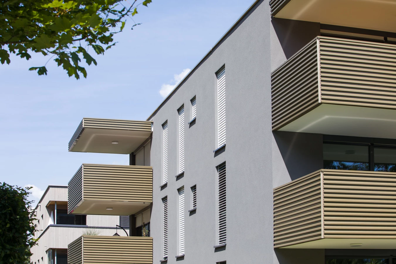 Graue Fassade der Wohnanlage Bahnhofstrasse mit mehreren Balkonen und eleganten Holzlatten-Brüstungen bei Tageslicht, umgeben von grünen Bäumen in Vandans (Bahnhofsstrasse)