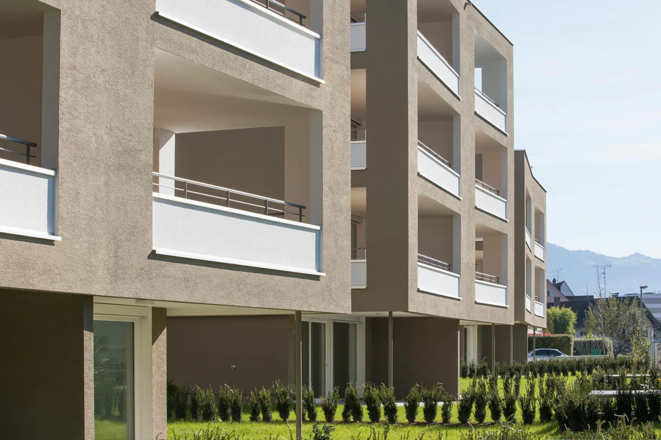 Moderne Wohnanlage Widum Lustenau mit beigefarbener Fassade, weißen Balkonen und grüner Gartenanlage unter blauem Himmel.