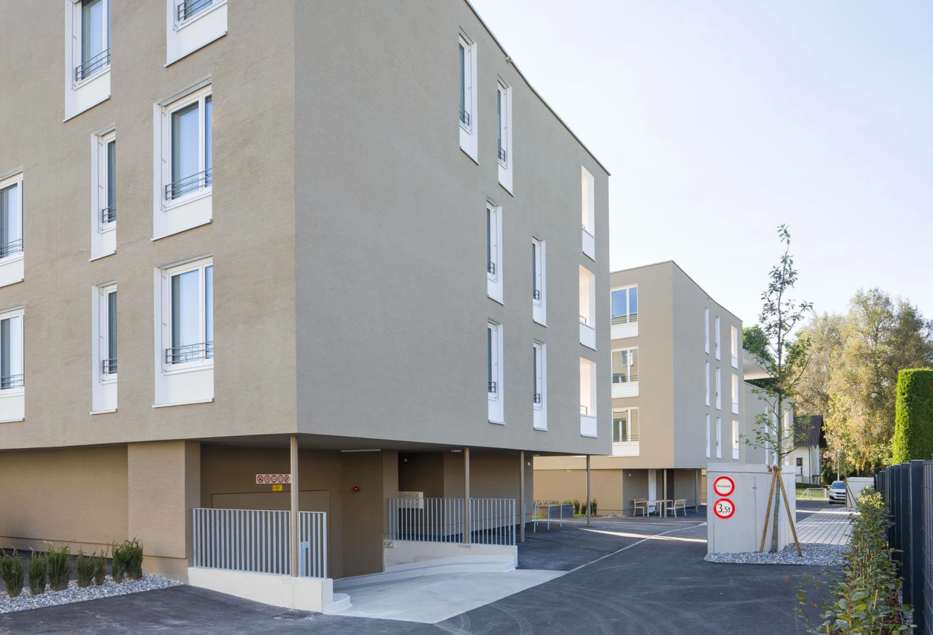 Moderne Wohnanlage Widum Lustenau 13 mit beige Fassaden, weiß umrahmten Fenstern, Tiefgarageneinfahrt und Straßenschildern.