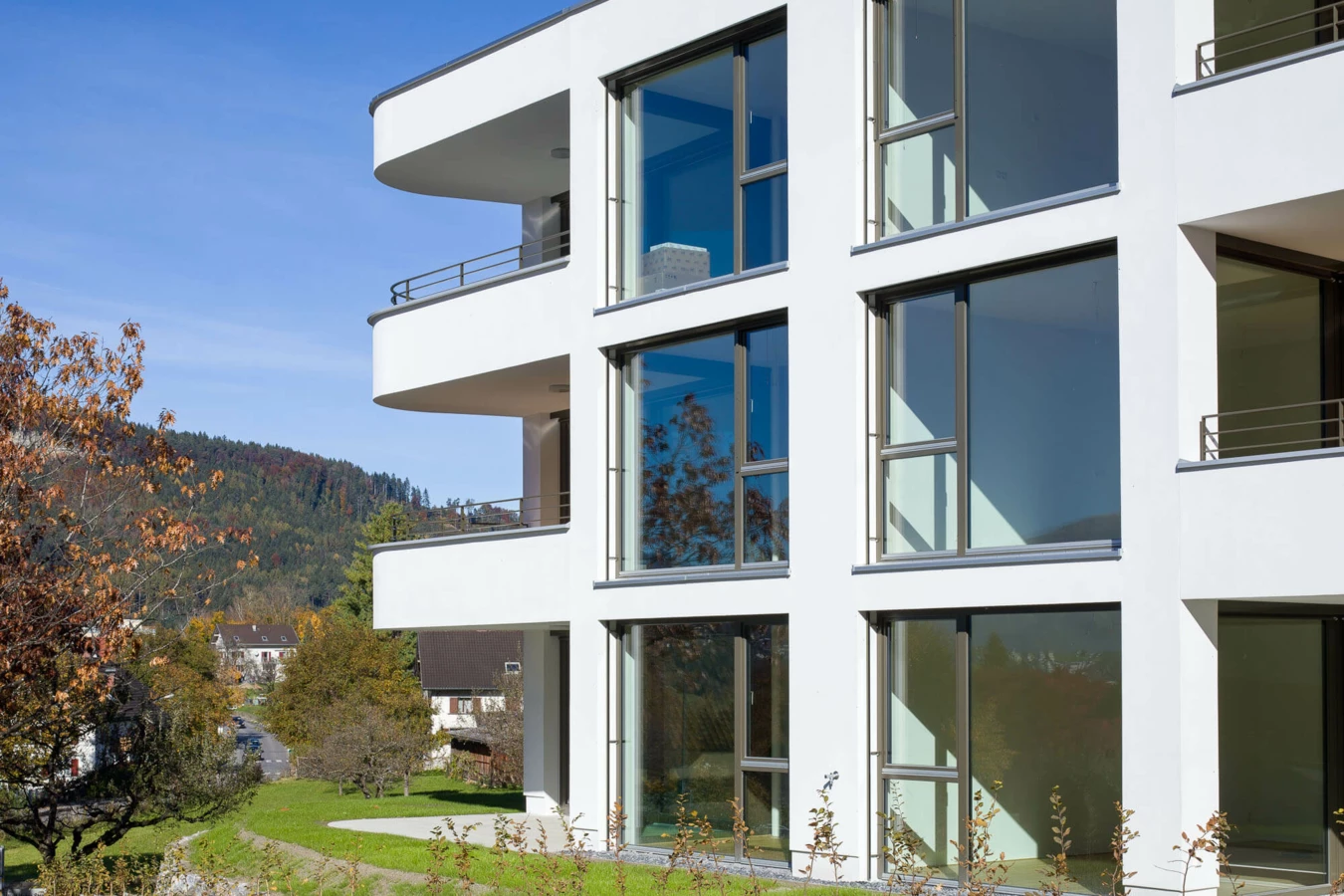Moderne, weiße Wohnanlage in Feldkirch mit großen Fenstern und Balkonen vor einem bewaldeten Hügel an einem sonnigen Tag (Wolf-Huber-Straße).