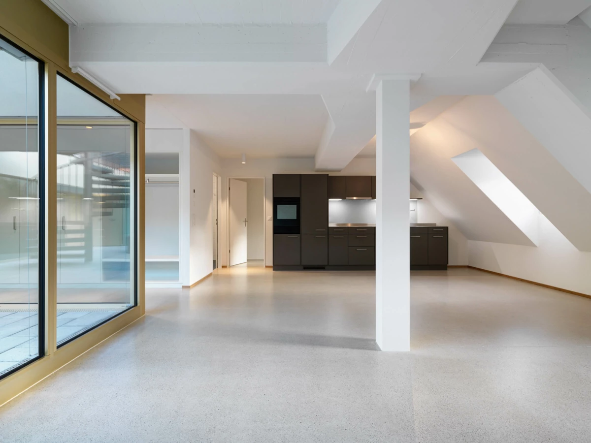 Eine moderne, leere Küche mit dunklen Schränken und einem Backofen in einem hellen Raum mit Dachschrägen und einem breiten Fenster (Multergasse, Gallen).
