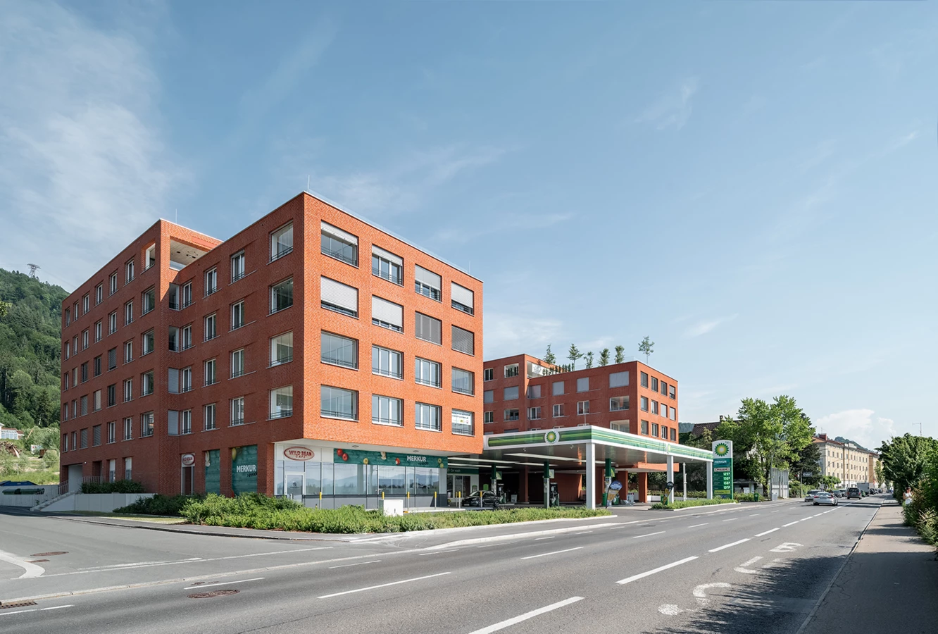 Zwei moderne Backsteingebäude neben einer BP-Tankstelle, mit "Merkur"- und "Wild Bean Cafe"-Beschriftungen, an einer Straße in Lochau (Seeblick).