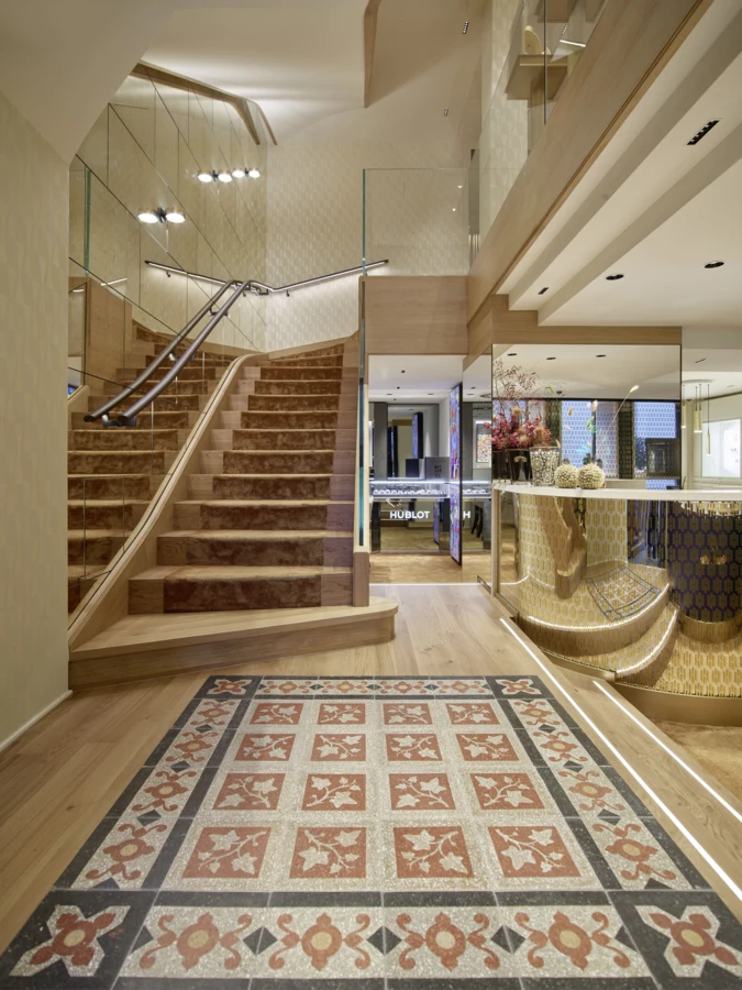 Geschäftsinterieur mit Holztreppe mit braunem Teppich, gemusterter Fliesenboden und beleuchteter Vitrine mit Hublot-Schriftzug (Huber, Rathausstr).