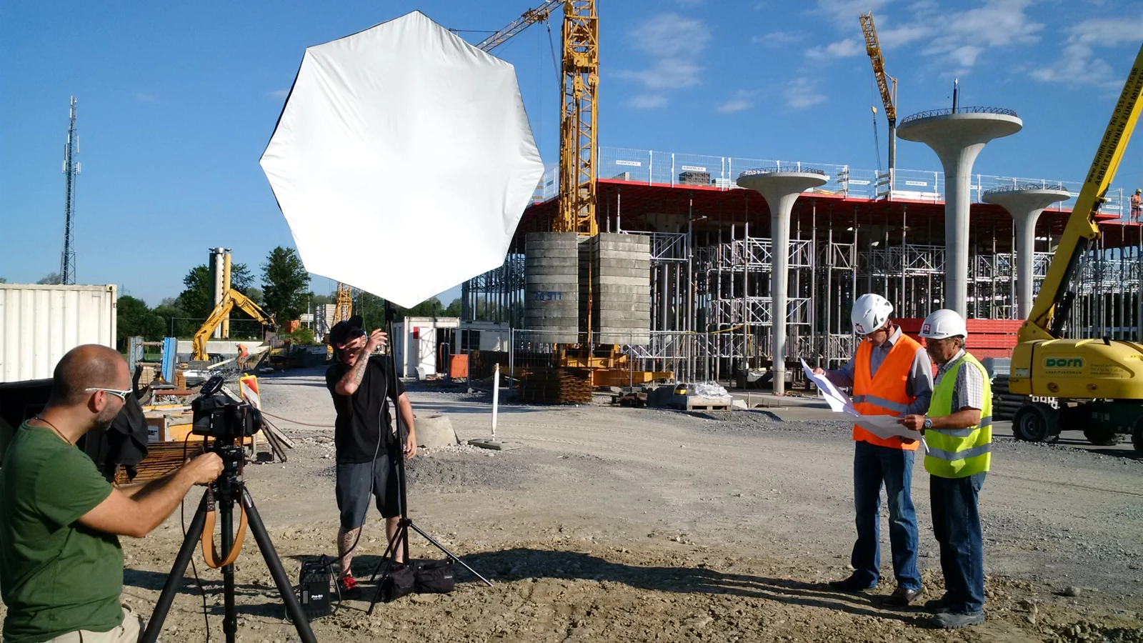 Fotoshooting auf einer Baustelle mit Kameramann, einem Lichttechniker und zwei Bauarbeitern, die Pläne begutachten.
