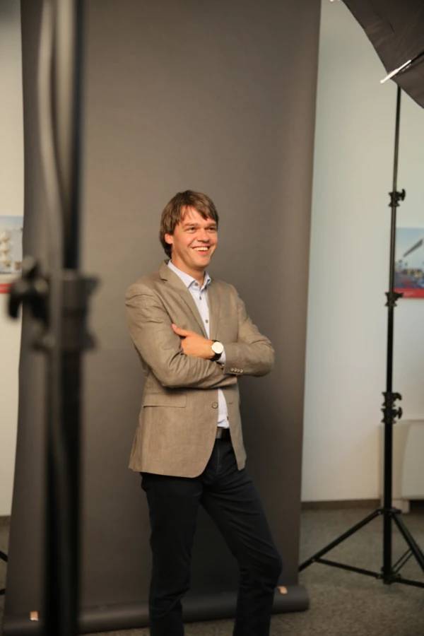 Ein lächelnder Mann in einem beigen Sakko und dunkler Hose steht mit verschränkten Armen vor einem grauen Hintergrund in einem Fotostudio.
