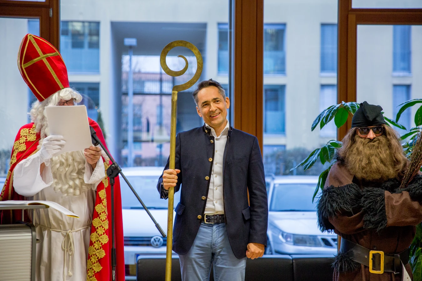Ein Mann im Trachtenjanker hält einen Bischofsstab, flankiert von "Nikolaus" und "Knecht Ruprecht" vor einem Gebäude in Dornbirn (Hubert, Geburtstag).