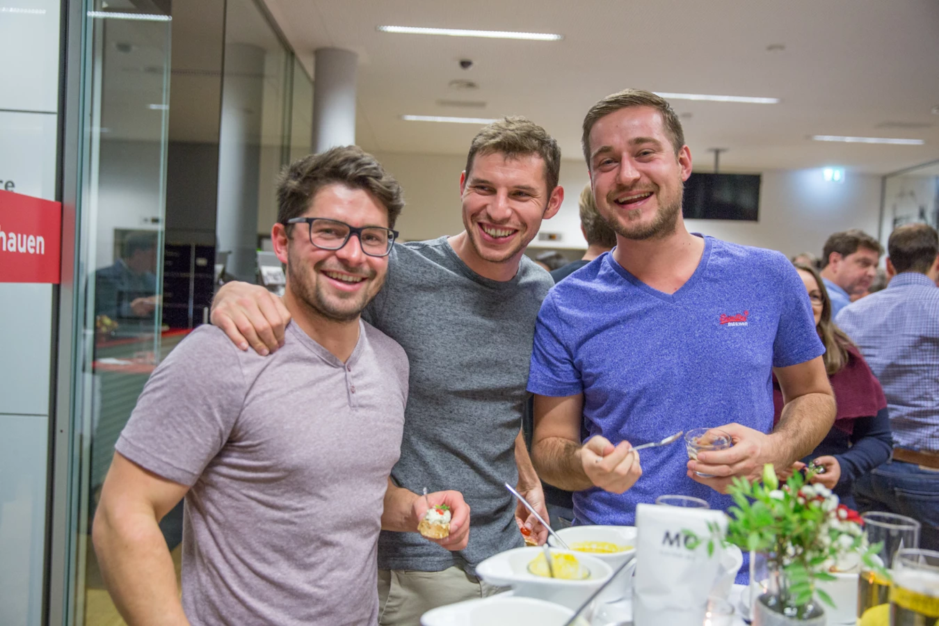 Drei lachende Männer stehen bei einer Veranstaltung vor einem Buffet mit kleinen Speisen und Getränken (Hubert, Geburtstag).