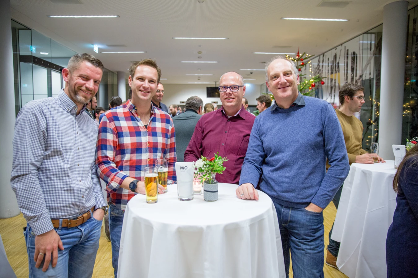 Vier lächelnde Männer unterhalten sich an einem Stehtisch mit Getränken bei einer Veranstaltung (Hubert, Geburtstag).