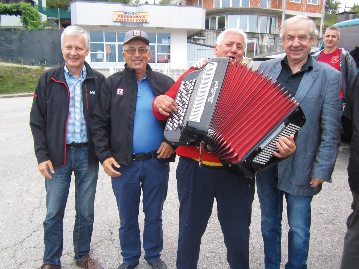 Four men standing outdoors, one playing a red and black Dallapè accordion, with buildings in the background (pensioners).