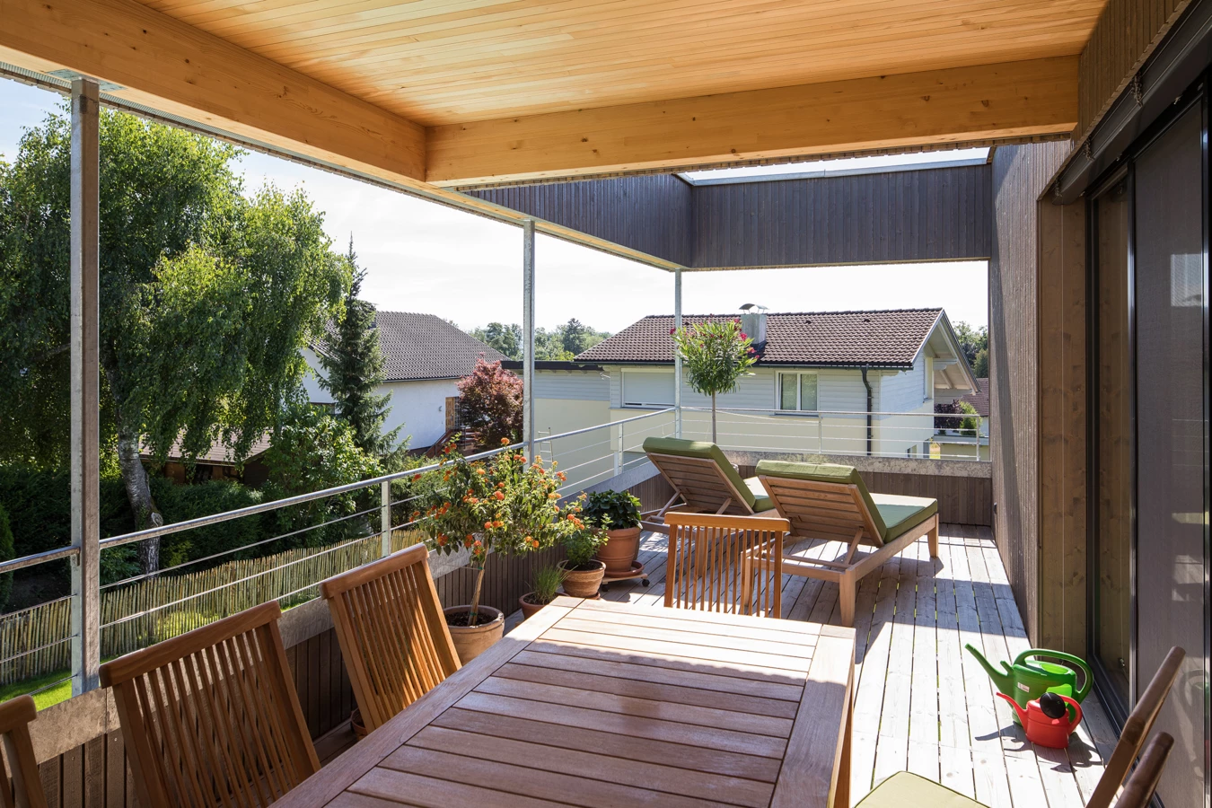 Eine große Holzterrasse mit Esstisch, Stühlen und Sonnenliegen unter einem geschützten Dach, umgeben von Bäumen und Häusern (Hohenweiler).