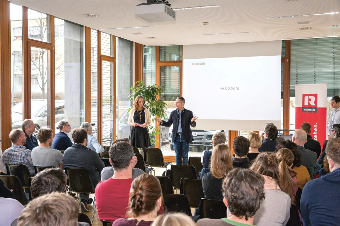Zwei Personen präsentieren vor einem Publikum, mit Rhomberg Bau Flagge und Sony Logo an der Wand (Digital, Day).