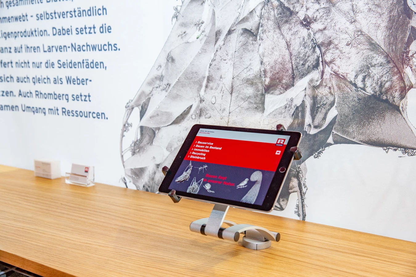 Tablet mit Informationen zu Rhomberg Bau Leistungen auf Holztheke vor einer Wand mit Blattmotiven und Text.