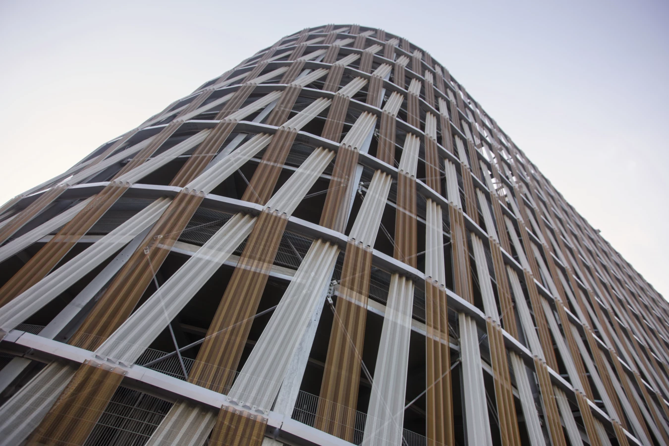 Ansicht der gebogenen Fassade eines Parkhauses mit vertikalen Lamellen in Braun- und Silbertönen unter hellem Himmel (Hochgarage, Wien, Hauptbahnhof).