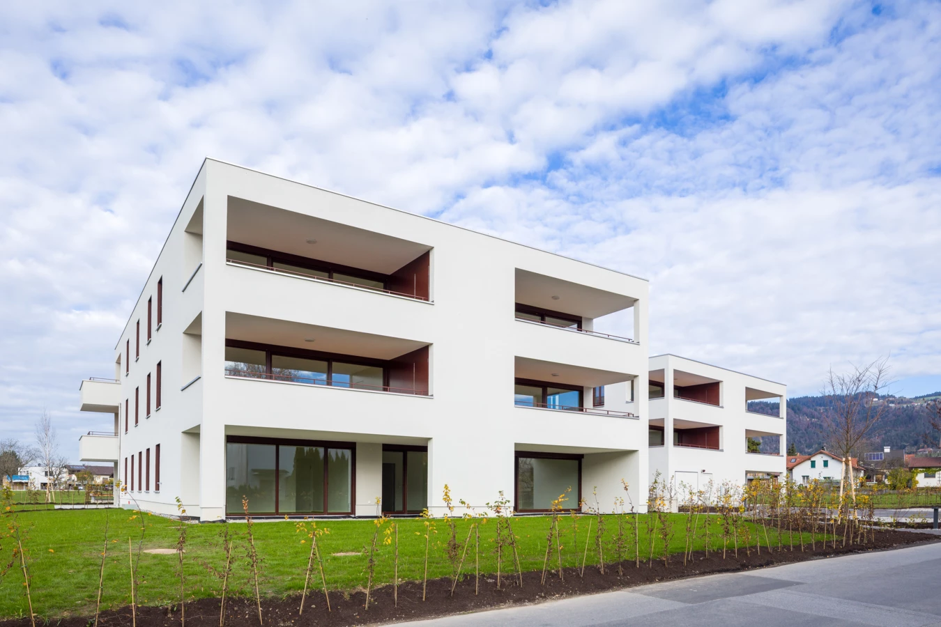 Zwei moderne weiße Wohngebäude mit Balkonen und großen Fenstern, umgeben von jungem Grün und Rasen, unter einem bewölkten Himmel (Unterhochstegstrasse).