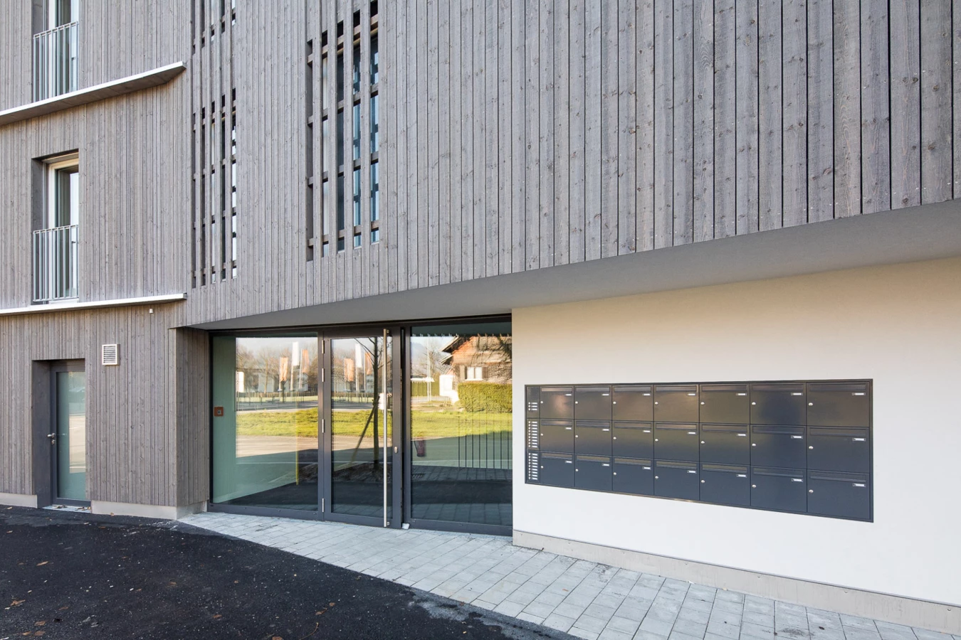 Eingangsbereich eines modernen Gebäudes mit Holzfassade, großen Glasfenstern und einer Reihe von Briefkästen an der weißen Wand (Froschweg).