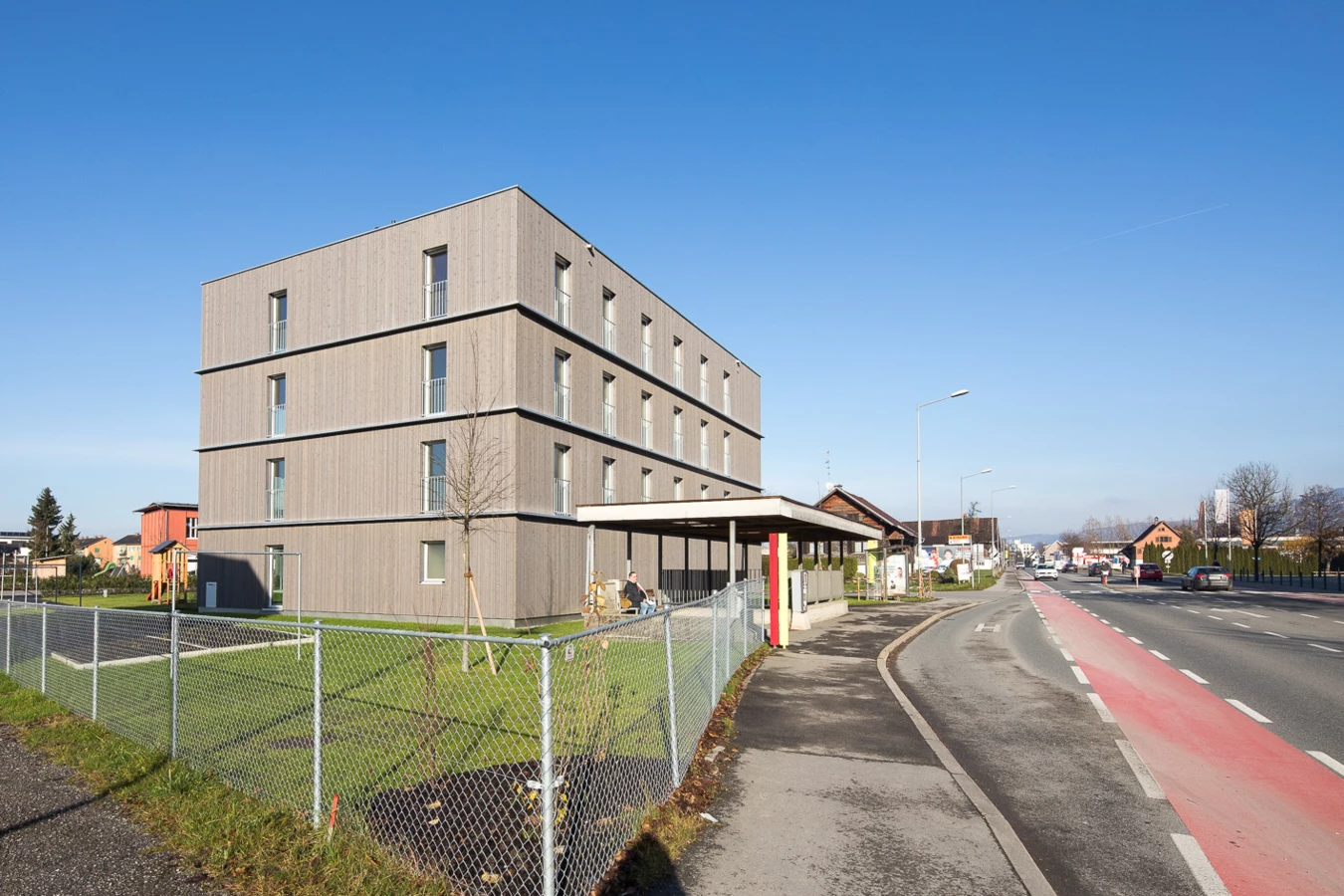 Modernes Mehrfamilienhaus mit Holzfassade und Balkonen am "Froschweg" in Dornbirn. Vor dem Gebäude befindet sich eine Parkgarage, ein Spielplatz und ein Gehsteig neben einer Straße.