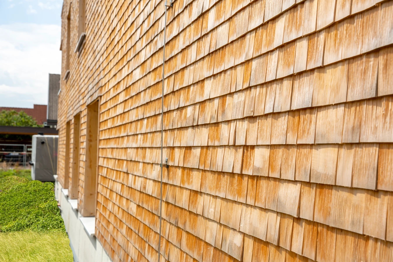 Seitenansicht eines modernen Gebäudes mit einer Fassade aus Holzschindeln, davor ein grüner Rasen unter blauem Himmel (Hellbrunnenstraße).