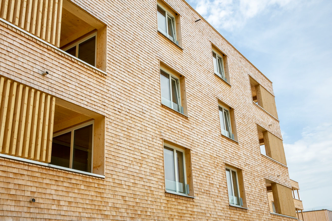 Fassadenansicht eines modernen Gebäudes mit Holzschindelverkleidung und großen Fenstern sowie hölzernen Lamellen an den Balkonen (Hellbrunnenstraße).