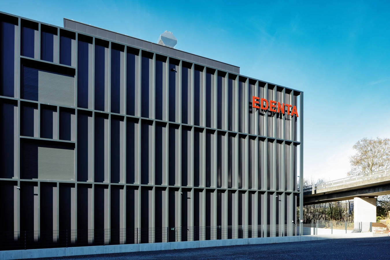 Modernes dunkelgraues Gebäude mit vertikalen Lamellen und dem roten Edenta-Logo an der Fassade unter blauem Himmel, daneben eine Brücke.