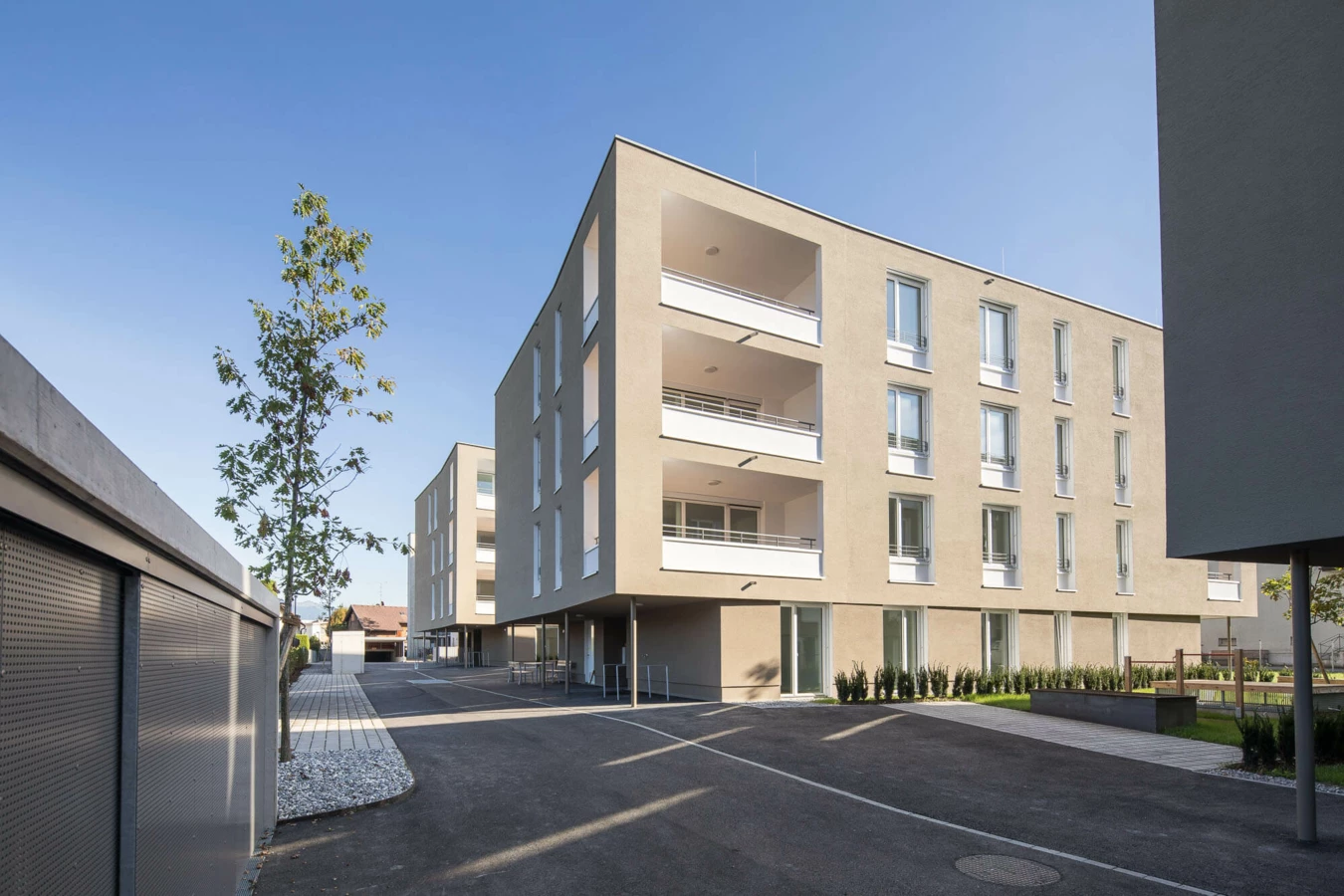 Wohnanlage Widum Lustenau 14 mit modernen Fassaden, Balkonen und bodentiefen Fenstern, umgeben von asphaltierten Wegen und minimalistischer Bepflanzung.