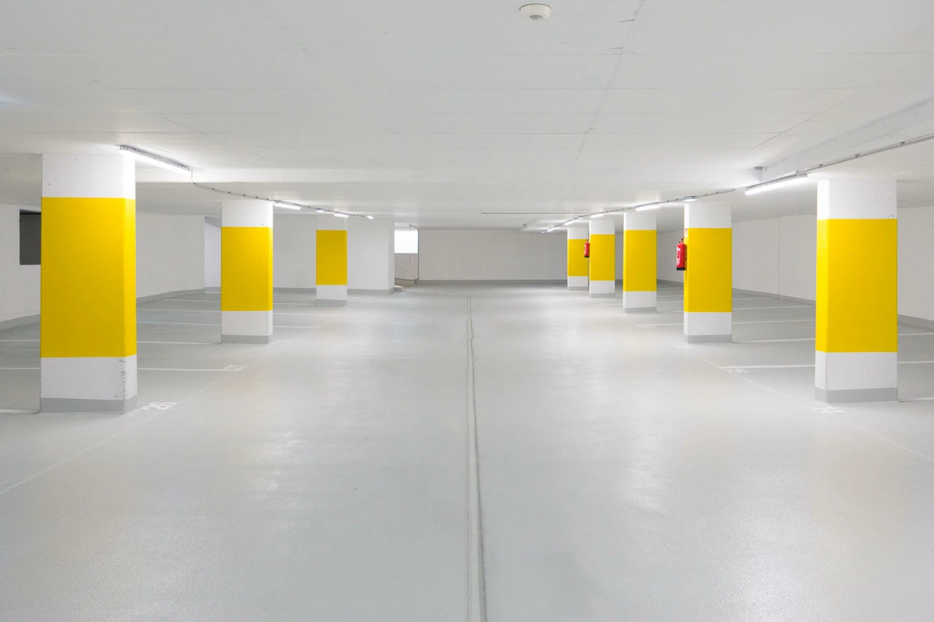 Leere Tiefgarage mit grauer Bodenfläche, weißen Wänden und gelb-weiß gestrichenen Säulenreihen mit Parkplatzmarkierungen (Widum, Wohnanlage, Lustenau).